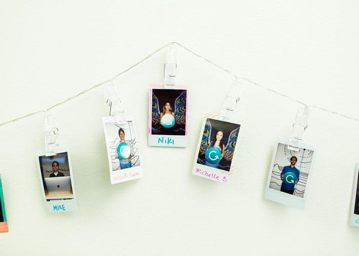 Five polaroid photos of smiling staff members hang on a wire against a blank wall. In the photos, the employees are holding a cutout of the Grammarly logo.