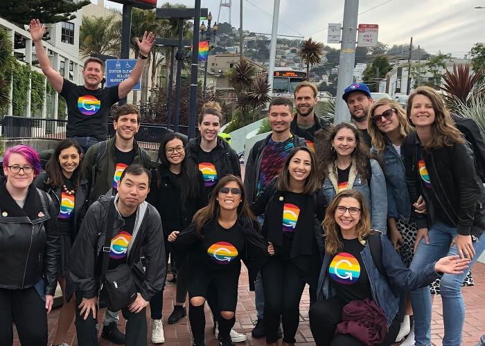 Sixteen people wearing matching shirts with a rainbow Grammarly logo pose on a brick sidewalk in San Francisco and smile at the camera. In the background, there are houses on a hill, trees, a bus, street signs, and a rainbow flag flying on a light pole.