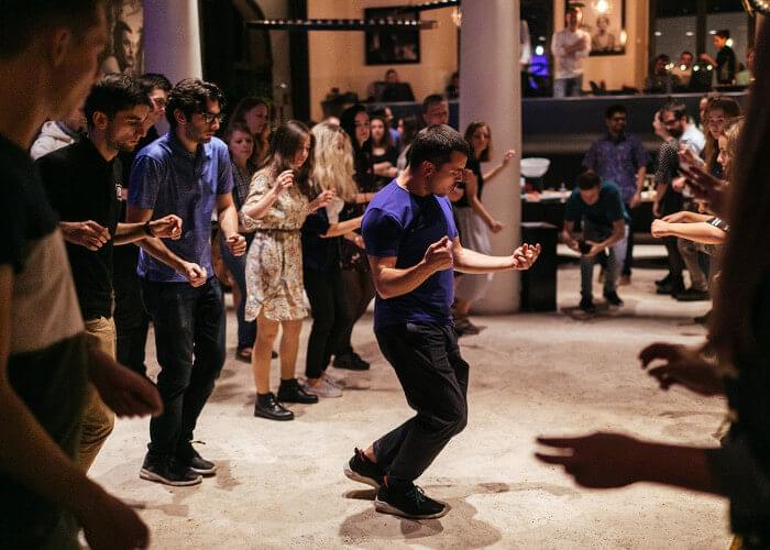 A diverse group of people dance in a large event space with ambient lighting. One person dances in the middle of the group while someone films and others watch in the background.